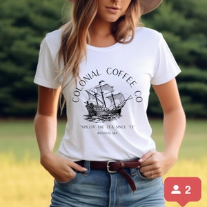 May include: White t-shirt featuring a black and white illustration of a tall ship with the text "COLONIAL COFFEE CO" and "Spillin' the Tea Since '73 Boston, MA". The shirt is paired with blue jeans and a brown belt.