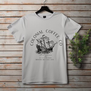 May include: A white t-shirt with a black graphic of a tall ship and the text "COLONIAL COFFEE CO". The text below reads "I LIKE MY COFFEE BLACK AND MY TEA IN THE HARBOR BOSTON, MA &bull; EST. 1773". The shirt is hanging on a black hanger.