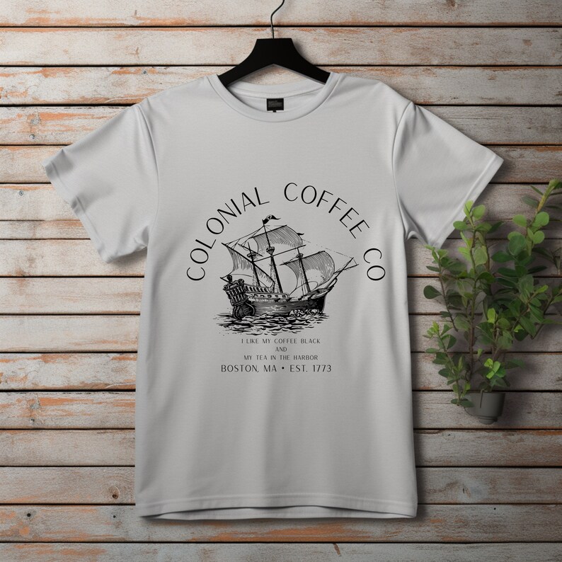May include: A white t-shirt with a black graphic of a tall ship and the text "COLONIAL COFFEE CO". The text below reads "I LIKE MY COFFEE BLACK AND MY TEA IN THE HARBOR BOSTON, MA &bull; EST. 1773". The shirt is hanging on a black hanger.