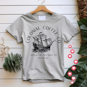 May include: A light gray t-shirt with the words "COLONIAL COFFEE CO" arched above a black and white illustration of a tall ship. Below the ship, the text reads "'SPILLIN' THE TEA SINCE '73" and "BOSTON, MA."