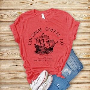 May include: Red t-shirt with a black graphic of a sailing ship and the text "COLONIAL COFFEE CO." and "I LIKE MY COFFEE BLACK AND MY TEA IN THE HARBOR BOSTON, MA EST. 1773." The shirt is paired with blue jeans and white sneakers.