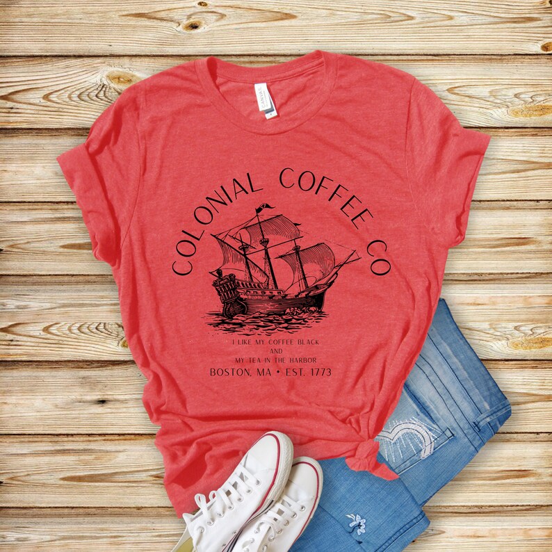 May include: Red t-shirt with a black graphic of a sailing ship and the text "COLONIAL COFFEE CO." and "I LIKE MY COFFEE BLACK AND MY TEA IN THE HARBOR BOSTON, MA EST. 1773." The shirt is paired with blue jeans and white sneakers.