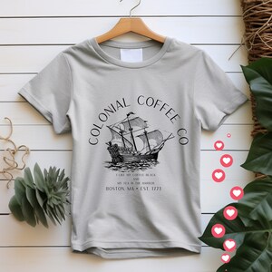 May include: A light gray t-shirt featuring a black and white illustration of a tall ship. The shirt has the text "COLONIAL COFFEE CO" in a semi-circle above the ship. Below the ship is the text "I LIKE MY COFFEE BLACK AND MY TEA IN THE HARBOR BOSTON, MA &bull; EST. 1773."