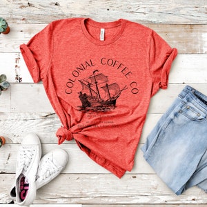 May include: Red t-shirt with the text "COLONIAL COFFEE CO" and a black illustration of a ship. The shirt also includes the text "LIKE ME COFFEE BLACK AND MY TEA IN THE HARBOR BOSTON MA EST 1773". White sneakers and blue jeans are next to the shirt.
