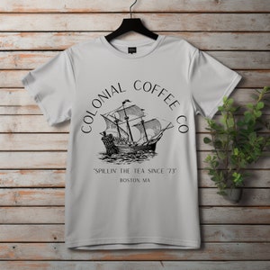 May include: A light gray t-shirt with a black graphic of a sailing ship and the words "COLONIAL COFFEE CO" in a circular design. Below the ship, the text reads "SPILLIN' THE TEA SINCE '73" and "BOSTON, MA". The shirt hangs on a black hanger.