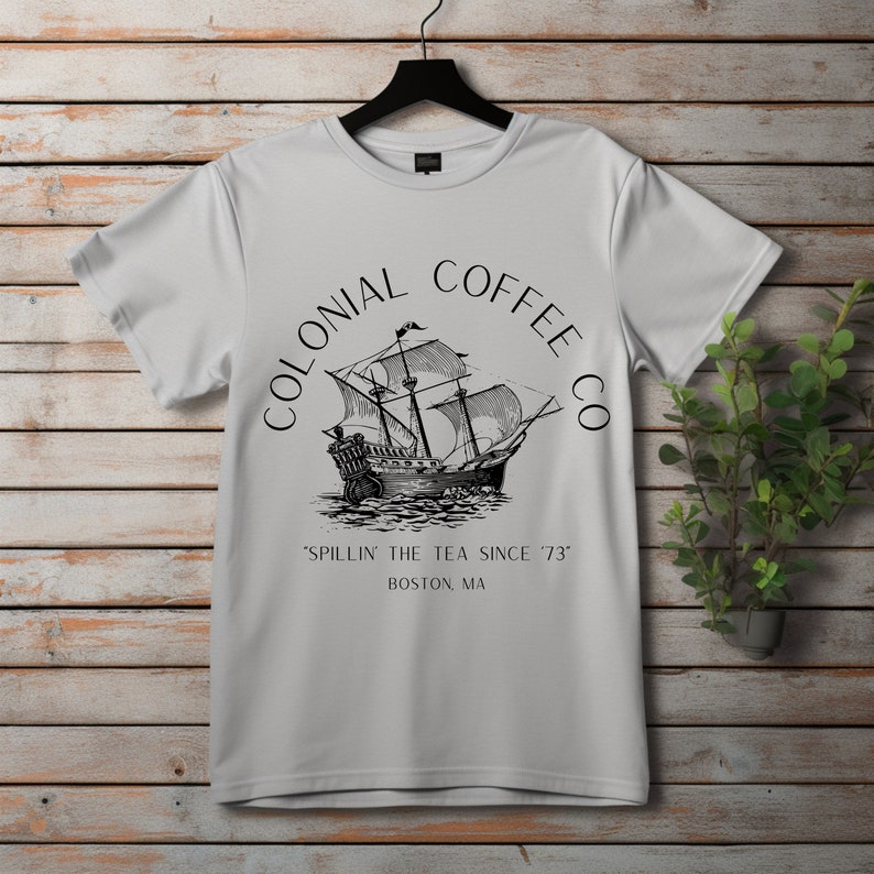 May include: A light gray t-shirt with a black graphic of a sailing ship and the words "COLONIAL COFFEE CO" in a circular design. Below the ship, the text reads "SPILLIN' THE TEA SINCE '73" and "BOSTON, MA". The shirt hangs on a black hanger.