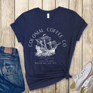 May include: Navy blue t-shirt with a white graphic of a sailing ship and the text "COLONIAL COFFEE CO." and "I LIKE MY COFFEE BLACK AND MY TEA IN THE HARBOR BOSTON, MA &bull; EST. 1773." The shirt is paired with blue jeans and white sneakers.