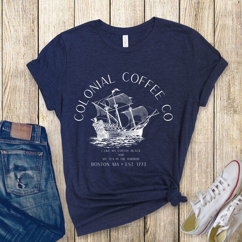 May include: Navy blue t-shirt with a white graphic of a sailing ship and the text "COLONIAL COFFEE CO." and "I LIKE MY COFFEE BLACK AND MY TEA IN THE HARBOR BOSTON, MA &bull; EST. 1773." The shirt is paired with blue jeans and white sneakers.