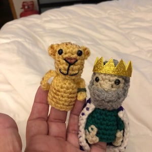 May include: Two crocheted finger puppets, one a yellow lion with black eyes and a pink nose, the other a gray-haired king with a gold crown and a green robe.