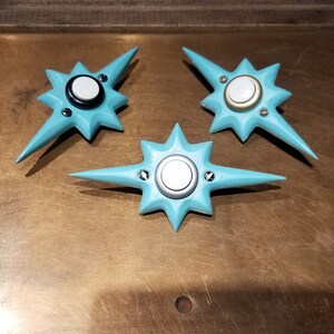 May include: Three turquoise star-shaped objects with a central circular design and pointed edges. Each has a silver or gold-colored center and is secured with small screws. The objects are arranged on a textured, brown surface.