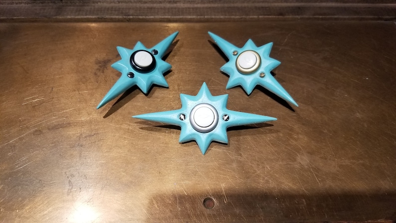 May include: Three turquoise star-shaped objects with a central circular design and pointed edges. Each has a silver or gold-colored center and is secured with small screws. The objects are arranged on a textured, brown surface.