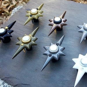 May include: A collection of starburst-shaped cabinet knobs in various metallic finishes, including black, bronze, silver, and gold. Each knob features a central white disc and is mounted on a black base with screws. The knobs are arranged on a dark gray surface.