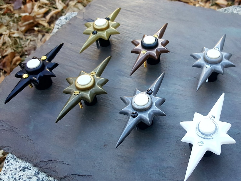 May include: A collection of starburst-shaped cabinet knobs in various metallic finishes, including black, bronze, silver, and gold. Each knob features a central white disc and is mounted on a black base with screws. The knobs are arranged on a dark gray surface.