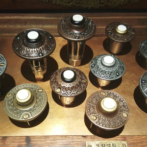 May include: A collection of antique doorbells with ornate, detailed designs. The doorbells feature a variety of finishes, including bronze, silver, and brass, with white push buttons. The collection is arranged on a wooden surface.