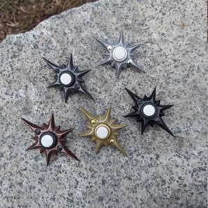 May include: Five star-shaped fidget spinners in various metallic finishes: black, silver, bronze, gold, and dark gray. Each spinner has a central button and pointed arms radiating outwards. The spinners are displayed on a textured stone surface.