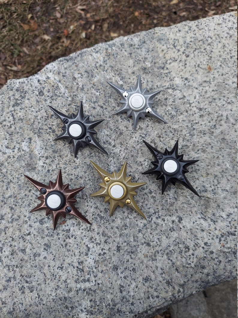 May include: Five star-shaped fidget spinners in various metallic finishes: black, silver, bronze, gold, and dark gray. Each spinner has a central button and pointed arms radiating outwards. The spinners are displayed on a textured stone surface.