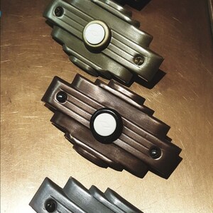 May include: Four Art Deco style doorbells in various colors: black, olive green, bronze, and gray. Each doorbell has a circular button with a white center and a decorative, stepped design. The doorbells are arranged on a brown surface.