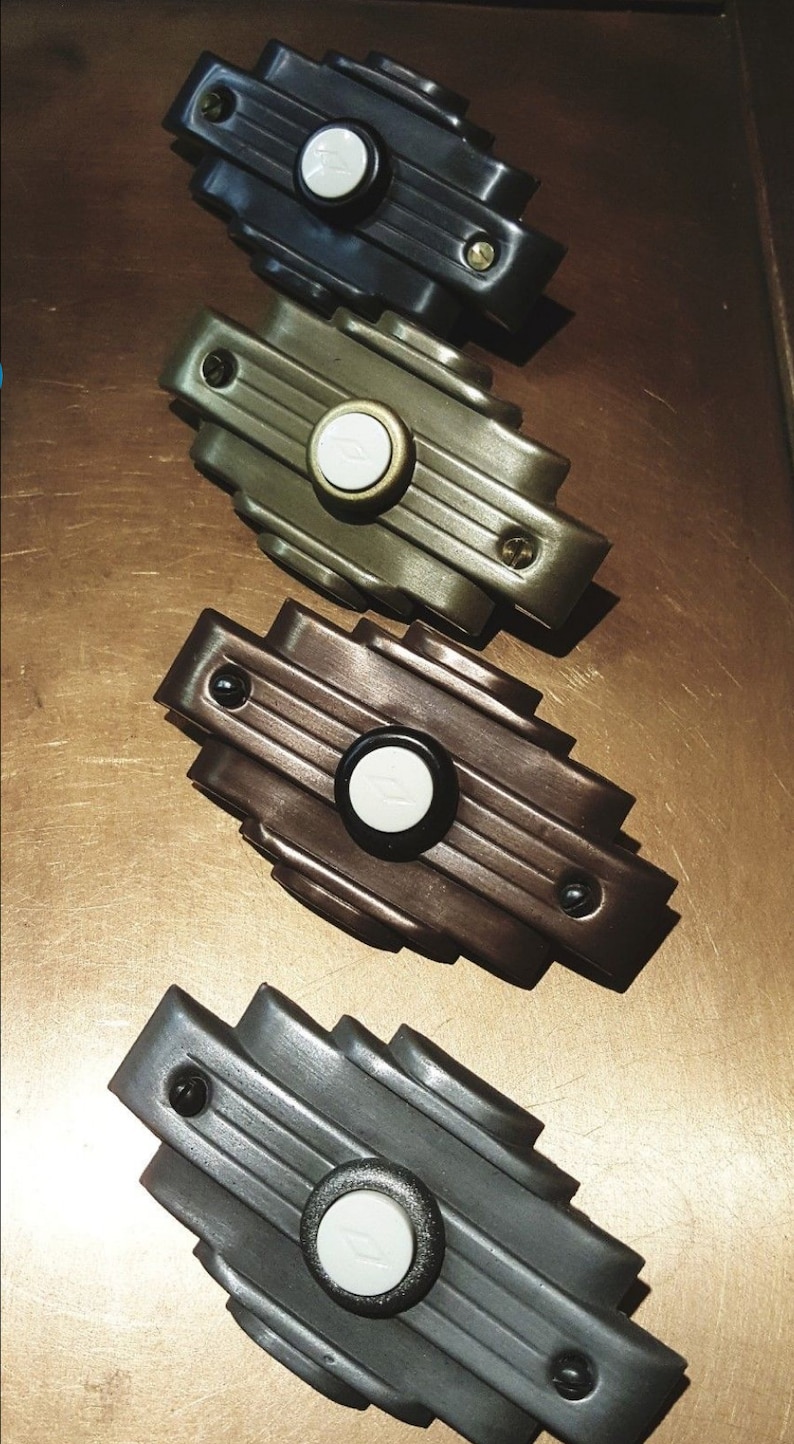 May include: Four Art Deco style doorbells in various colors: black, olive green, bronze, and gray. Each doorbell has a circular button with a white center and a decorative, stepped design. The doorbells are arranged on a brown surface.