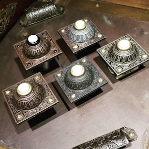 May include: An assortment of ornate, square doorbell buttons in various metallic finishes, including bronze, silver, and black. Each button features a central, circular push-button with a light-colored center and is accented with decorative screws. The buttons are arranged on a dark surface.