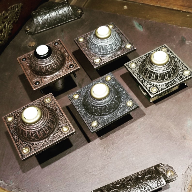May include: An assortment of ornate, square doorbell buttons in various metallic finishes, including bronze, silver, and black. Each button features a central, circular push-button with a light-colored center and is accented with decorative screws. The buttons are arranged on a dark surface.