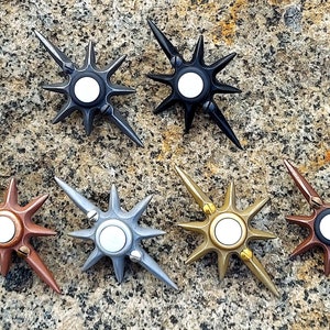 May include: Six doorbells in different finishes, including black, silver, copper, and gold. Each doorbell has a starburst design with a white button in the center.