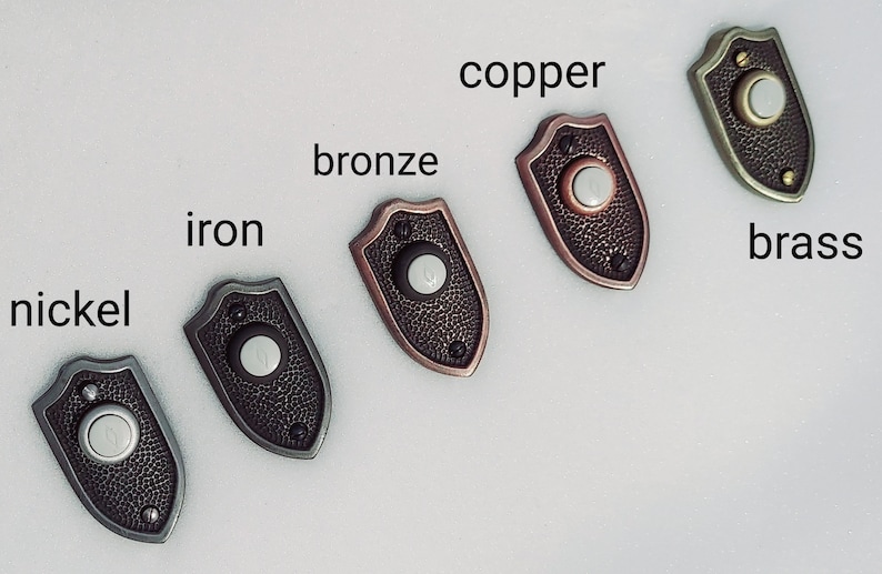 May include: Five decorative doorbell buttons in nickel, iron, bronze, copper, and brass. Each button has a shield shape with a textured surface and a circular button in the center. The names of the finishes are labeled on the image.