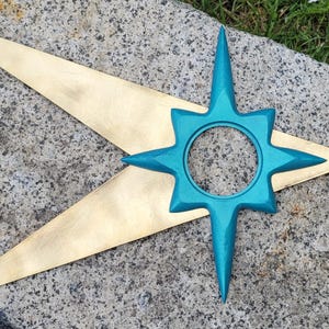 May include: A decorative star-shaped object featuring a large, flat, gold-colored metallic base with two long, pointed arms extending to the left. A smaller, bright teal, eight-pointed star with a circular cutout in the center is layered on top.
