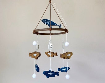 May include: A wooden baby mobile with a blue and white fish design. The mobile has a circular wooden frame with a blue fish hanging from the top. There are five smaller fish hanging from the frame, alternating between blue and brown. The mobile also has white pom-poms hanging from the frame. The text "SEA BEE MARKET" is printed on the frame.