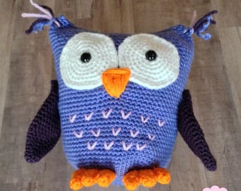 May include: A handmade crochet owl in shades of purple, white, orange, and black. The owl has large white eyes with black pupils, an orange beak, and orange feet. The wings and accents are dark purple. The owl is a soft, plush toy.