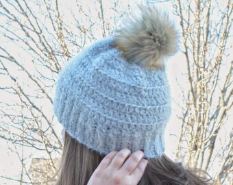 May include: A light grey knitted beanie hat with a fluffy, brown faux fur pom-pom. The hat has a ribbed design and a folded brim. The person in the image is wearing a dark green jumper. Bare tree branches are in the background.