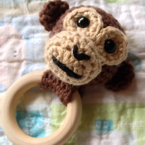 May include: A handmade crochet monkey baby rattle. The monkey has a light brown face, dark brown ears and head, and black eyes and mouth. The rattle features a natural wooden ring, perfect for little hands.