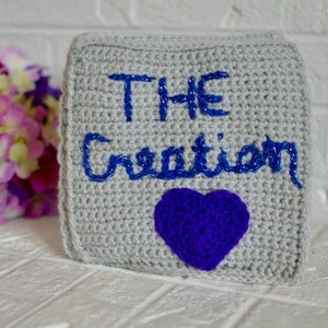 Puede incluir: Un cuadrado de crochet gris con las palabras "THE Creation" bordadas en azul. Un corazón de crochet morado se encuentra en la esquina inferior derecha.