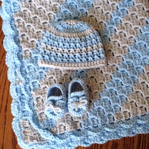 Könnte beinhalten: Eine blaue und graue gehäkelte Babydecke mit passender Mütze und Booties. Die Decke hat einen gewellten Rand.