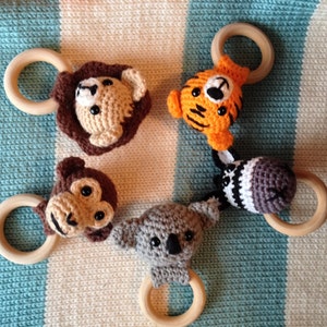 May include: Five handmade crochet baby toys featuring animal faces: a lion, tiger, monkey, koala, and zebra. Each toy has a wooden ring for easy gripping. The toys are in shades of brown, orange, gray, and white.