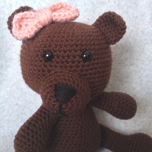 May include: A brown crocheted teddy bear with black eyes and a pink bow. The bear is sitting on a white surface.
