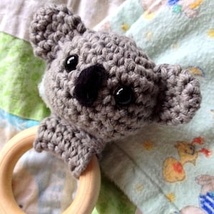 Op de afbeelding: Een grijs gehaakt koala-speelgoed bevestigd aan een houten bijtring. Het speelgoed heeft zwarte ogen en een zwarte neus.
