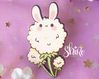 May include: A white wooden bunny rabbit pin with a fluffy dandelion design. The pin is on a pink background with white flowers and gold stars.