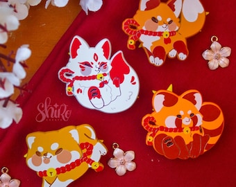 May include: Four enamel pins featuring cartoon foxes and a Shiba Inu dog, each with a red and gold color scheme. The pins are on a red background, with two flower charms and white flower blossoms in the background.