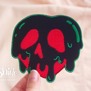 Pode incluir: Um remendo bordado em forma de maçã pingando com um rosto de caveira. A maçã é preta e vermelha com um contorno verde.