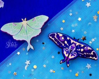 May include: Two wooden moth brooches, one green with white markings and one purple with black markings, on a blue tulle background with silver glitter stars.