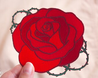 May include: A red felt rose with green felt stems and thorns. The rose is embroidered with a detailed design.