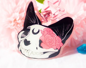 May include: A clear acrylic keychain featuring a black and white illustration of a cat skull with pink roses and a crescent moon. The keychain has a silver star charm.