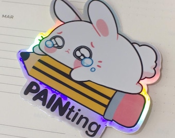 May include: A holographic sticker featuring a crying white bunny hugging a yellow pencil with the word "PAINTing" on it. The sticker is on an open planner with a blue marker and white flowers in the background.