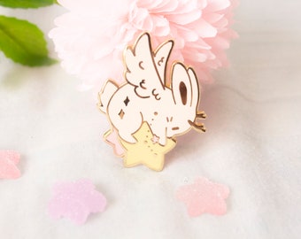 May include: A gold-colored enamel pin featuring a cream-colored bunny with angel wings, resting on a yellow star. The bunny has diamond-shaped accents. The pin is set against a soft pink flower and scattered star-shaped confetti.