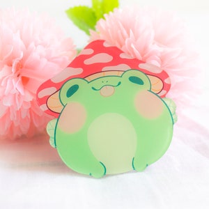 May include: A green frog wearing a red and white mushroom hat. The frog has a happy expression and is looking up. The frog is made of clear acrylic.