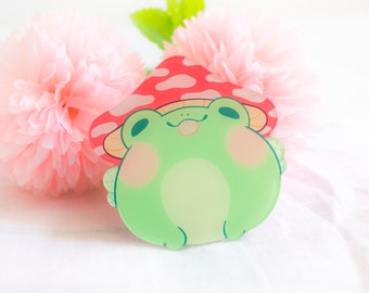 May include: A green frog wearing a red and white mushroom hat. The frog has a happy expression and is looking up. The frog is made of clear acrylic.