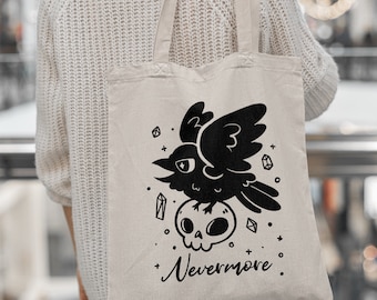 May include: A white canvas tote bag with black handles. The bag features a black and white illustration of a raven perched on a skull with the text "Nevermore."