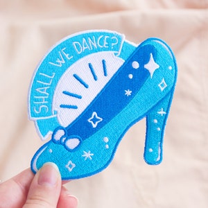 Può includere: Una toppa da stirare ricamata blu con una scarpetta di vetro con un fiocco bianco e il testo "Shall We Dance?"