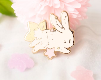 May include: A gold-colored bunny pin with a cream-colored body and star accents. The pin features a star-shaped bow and a pink star at the bottom. The pin is set against a soft pink floral background.