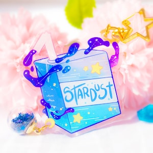 May include: A blue and purple galaxy-themed juice box keychain with a straw and the word "Stardust" printed on it. The keychain is made of clear acrylic and has a gold star charm attached.